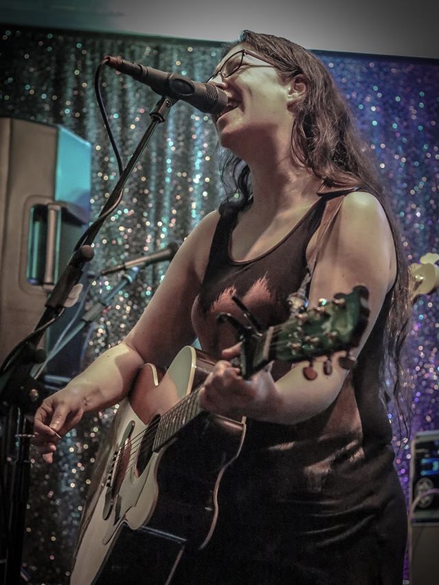 IndieFly singer/guitaist Emma Thorbinson singing at a show with long hair, glasses and a black tank top with a band logo