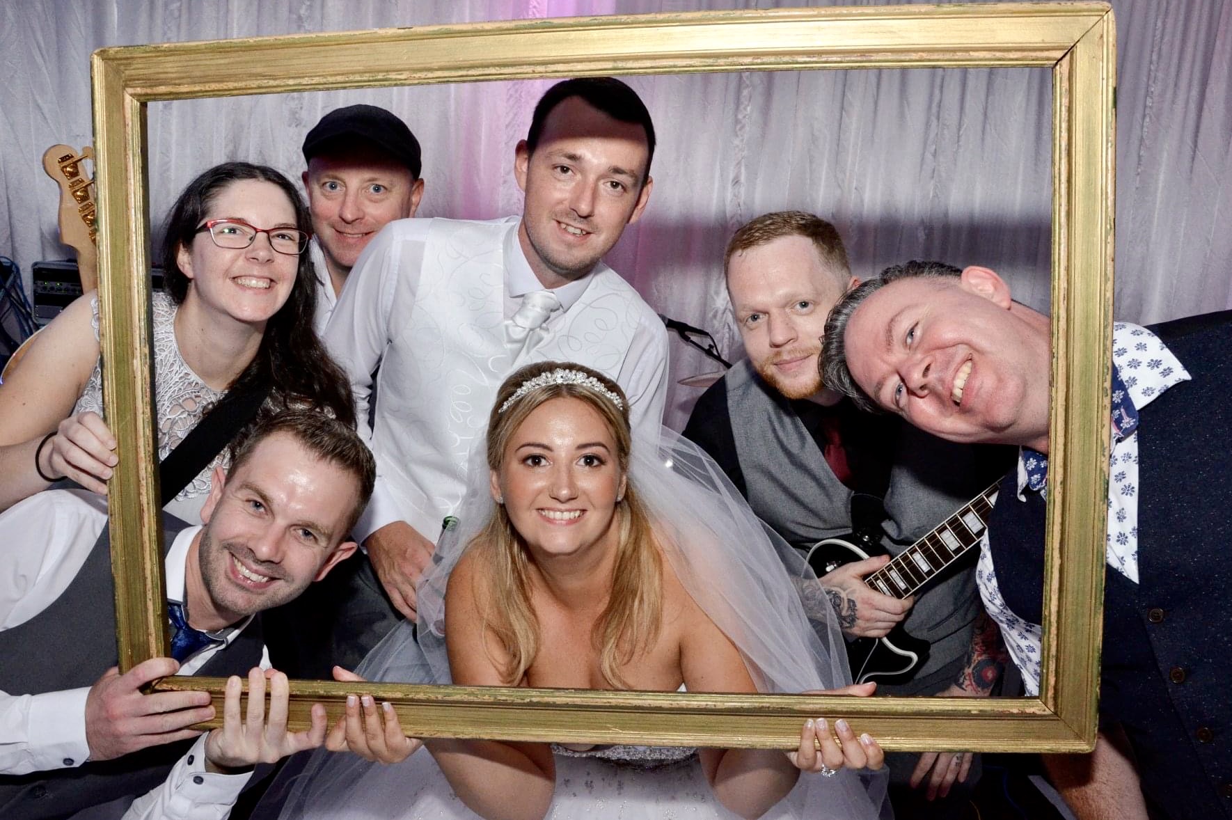 All smiles: IndieFly band hold a very large wooden pricture frame that frames a beautiful blonde bride in her wedding dress next to her groom. The band lean into the frame all around them.